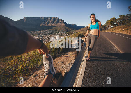 Fit young woman standing with man stretching his leg outdoors on country road. Young runners relaxing after morning run. POV sho Stock Photo