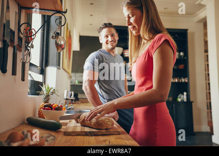 Indoor shot of young man and woman in kitchen during morning. Focus on woman cutting bread, preparing breakfast. Stock Photo