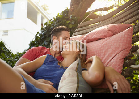 Young couple is cuddling on a hammock. Young man kissing forehead of his girlfriend smiling. Romantic couple outdoors relaxing o Stock Photo