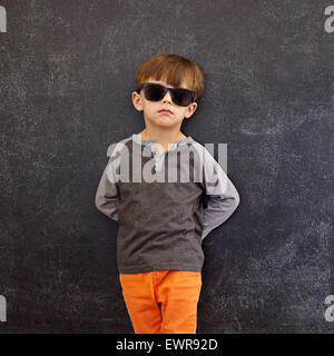 Cute little kid standing in front of the blackboard. Stylish little boy wearing sunglasses leaning on a blackboard looking at ca Stock Photo