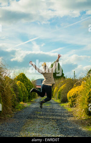 Happy Girl, woman, lady, female in a coat leaping in the air jumping with hands up on a country lane, blue sky and green bushes Stock Photo