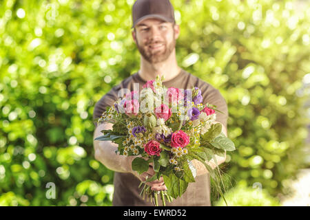 Three quarter length front view of smiling bearded 20s man wearing brown baseball cap, brown t-shirt and jeans holding bunch of Stock Photo