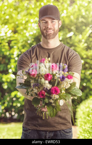 Three quarter length front view of smiling bearded 20s man wearing brown baseball cap, brown t-shirt and jeans holding bunch of Stock Photo