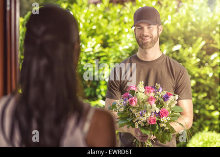 Smiling bearded 20s man wearing brown cap and brown t-shirt delivers flowers to door of young brunette female. Over the shoulder Stock Photo