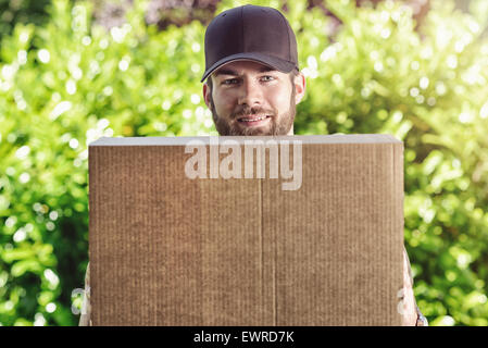 Smiling postman carrying a large brown cardboard parcel waiting to make a delivery at a house as he looks over the top of the bo Stock Photo