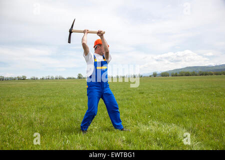 Worker with pick axe is digging on a wide green field. Stock Photo