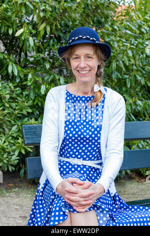 Caucasian middle aged woman in old-fashioned clothes with blue hat sitting on bench in park Stock Photo