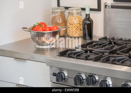 Colander containing ripe tomatoes, a bottle of olive oil, and a jar of rotini next to a stainless steel gas stove in kitchen Stock Photo