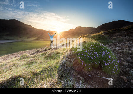 Man wearing blue shell jacket celebrating the sunset after a mountain hike, sun is setting behind a mountain range, and the last Stock Photo