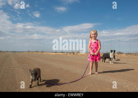 Keetmanshoop, Namibia, Africa - Young girl with small pig on a leash and dog behind her in the middle of a dirt road Stock Photo