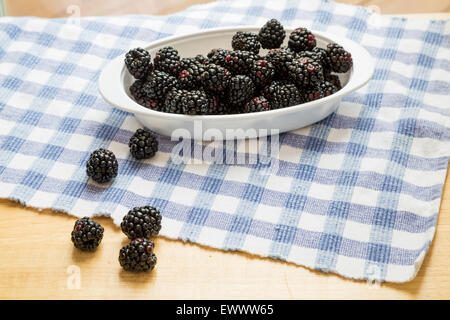 A white bowl of blackberries on a blue check placemat and wood table in window light Stock Photo