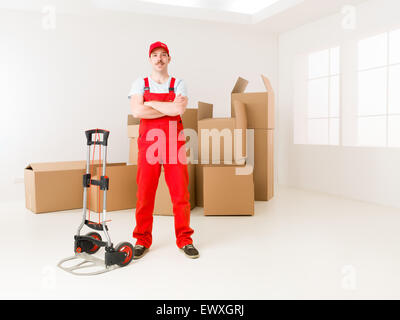 young confident delivery man standing in new home, holding hands crossed against his chest, with cardboard boxes in background Stock Photo