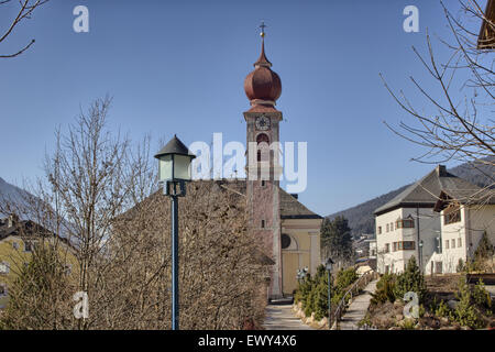 The Luis Trenker Promenade leading to Ortisei, Italian village in Dolomites Alps: typical homes, buildings and the red belfry with clock of St. Ulrich parish church with snowy mountains and green conifers in the background Stock Photo