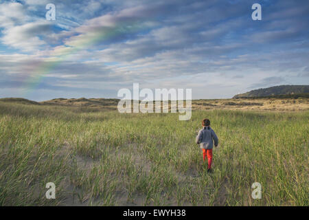 Rear view of a boy running through stand dunes with rainbow in the sky Stock Photo