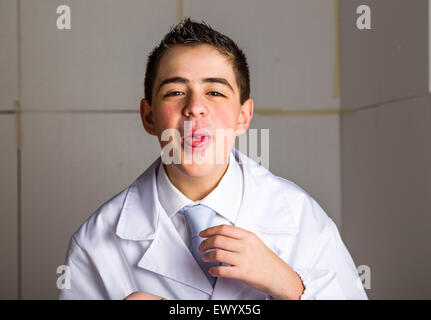 Boy dressed as a doctor in light blue tie and white coat helps to feel medicine more friendly: he is showing his tongue with a pill on it. His acne skin has not ben retouched Stock Photo