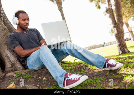 Young man leaning against tree, wearing headphones and using laptop Stock Photo