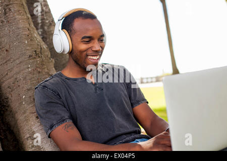 Young man leaning against tree, wearing headphones and using laptop Stock Photo