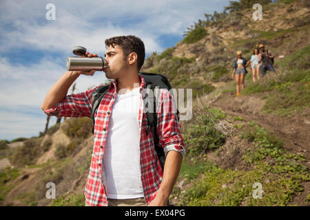 Young man at bottom of hill, drinking from flask, friends trailing behind Stock Photo