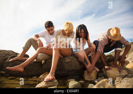 Group of friends sitting together on rocks on beach, looking in rock pools Stock Photo