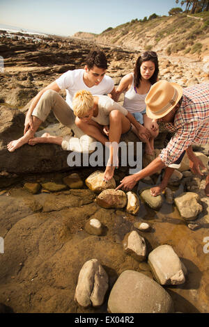 Group of friends sitting together on rocks on beach, looking in rock pools Stock Photo