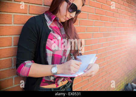 Young woman leaning against brick wall writing in notebook Stock Photo