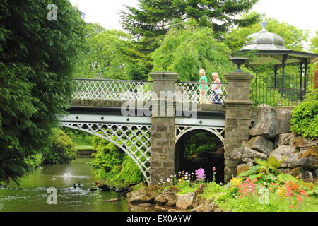 Women walk across a bridge near the bandstand (pictured) in the Pavilion Gardens in Buxton Derbyshire England - summer Stock Photo