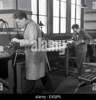 Historical, 1950s, at a further education technical college, inside a brightly room, a male student in overalls standing at a workbench using a metal vice, with an older man in a jacket and trousers, perhaps a teacher, checking a piece of metalwork, England, UK. Such colleges were organised on regional, area and local levels and funded by local authorities. Stock Photo