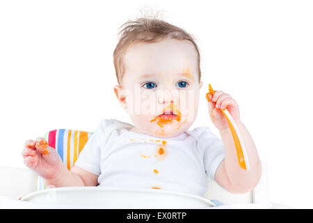 Funny messy baby eating her first solid vegetable food in a white high chair, isolated on white Stock Photo