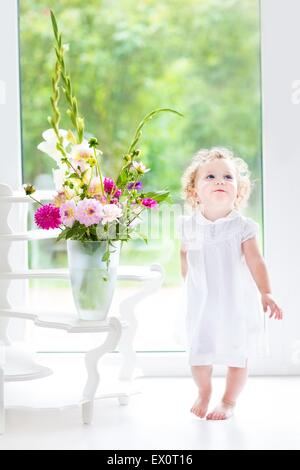 Beautiful curly baby girl in a white dress playing with a fresh flower bouquet next to a big window and door to the garden Stock Photo