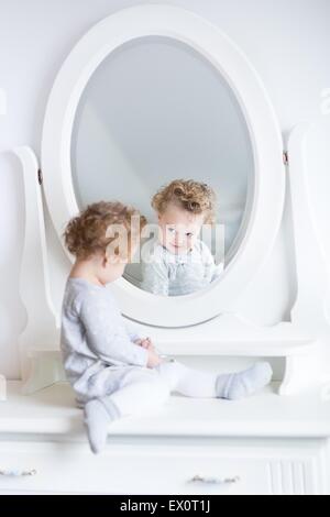 Funny cute baby girl watching her reflection in a white bedroom with a beautiful round mirror Stock Photo