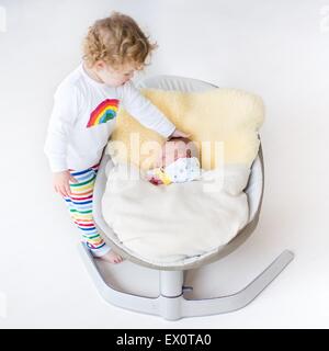 Tiny newborn baby sleeping in a swing on a sheepskin with his toddler sister standing next to him Stock Photo