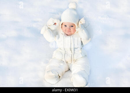 Funny little baby sitting in fresh snow wearing a white jacket and a warm knitted hat Stock Photo