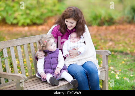 Young mother relaxing on a wooden bench in a colorful autumn park with her toddler daughter and newborn son