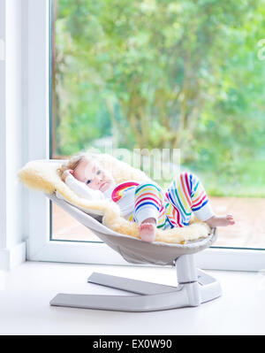 Cute little girl relaxing in a swing next to a big window Stock Photo