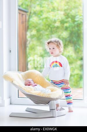 Cute toddler girl playing with her newborn baby brother relaxing in a swing next to a big window and door to the garden Stock Photo