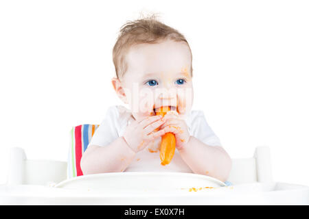 Sweet baby eating her first solid food in a white chair, isolated on white Stock Photo