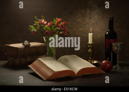 still life of an open bible dictionary on a table with a candle, wine bottle and goblet, apple and pocket watch Stock Photo