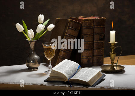 Still life image of an open bible on a table with old books, tulips in a vase, a glass of wine and a candle Stock Photo