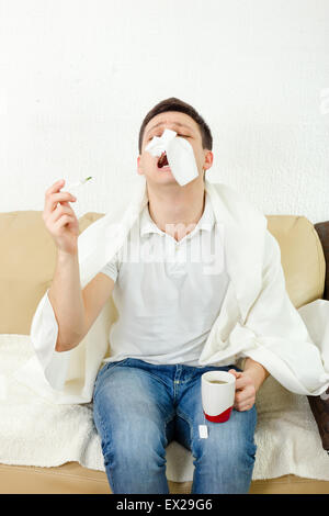 Young adult man suffering flu. Holding thermometer and tea in hand and has tissue paper on his nose, sneezing. Looking sad and d Stock Photo