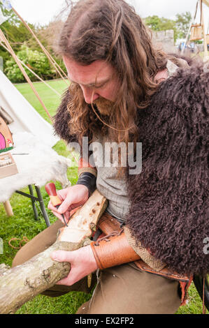 A man dressed in Iron Age clothing whittles a face into a piece of wood Stock Photo