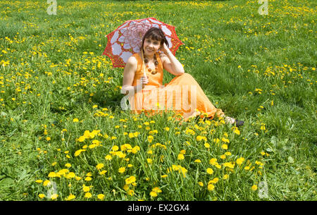 Pretty woman European brunetter in orange dress with white-red parasol sitting on meading with yelow dandelions. Stock Photo