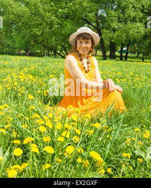European lady in hat and orange dress sitting on meadow with yellow dandelions. Stock Photo