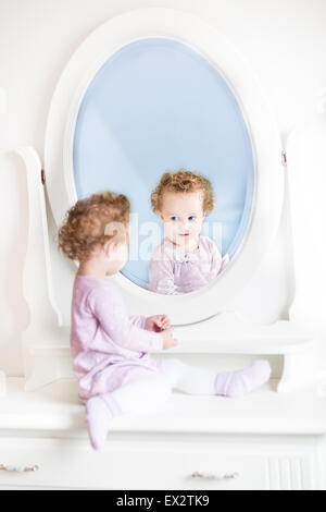 Cute little toddler girl with curly hair looking at her reflection in a beautiful white mirror Stock Photo