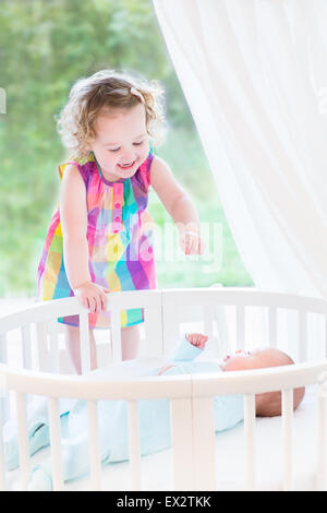 Cute laughing toddler girl playing with her newborn baby brother laying in white round crib next to big window with garden view Stock Photo