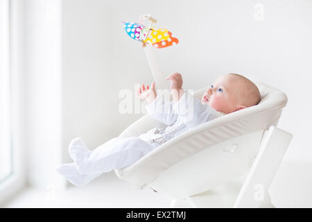 Cute newborn baby boy watching a colorful mobile toy sitting in a white high chair next to a window Stock Photo