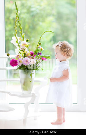 Beautiful toddler girl watching flowers in a big vase next to a window with garden view Stock Photo