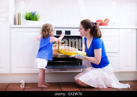 Mother and child bake a pie. Young woman and her daughter cook in a white kitchen. Kids baking pastry. Children helping to make Stock Photo