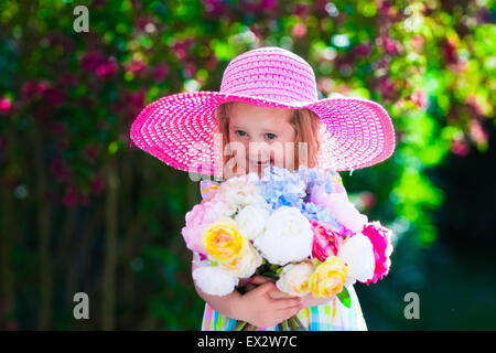 Little cute girl with flowers. Child wearing a pink hat playing in a blooming summer garden. Kids gardening. Stock Photo