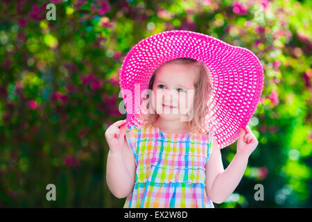 Little cute girl with flowers. Child wearing a pink hat playing in a blooming summer garden. Kids gardening. Stock Photo