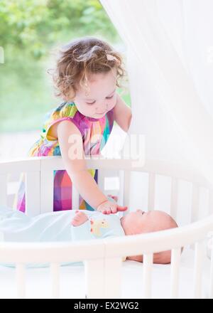 Cute toddler girl playing with her newborn baby brother laying in a white round crib next to a big window with garden view Stock Photo